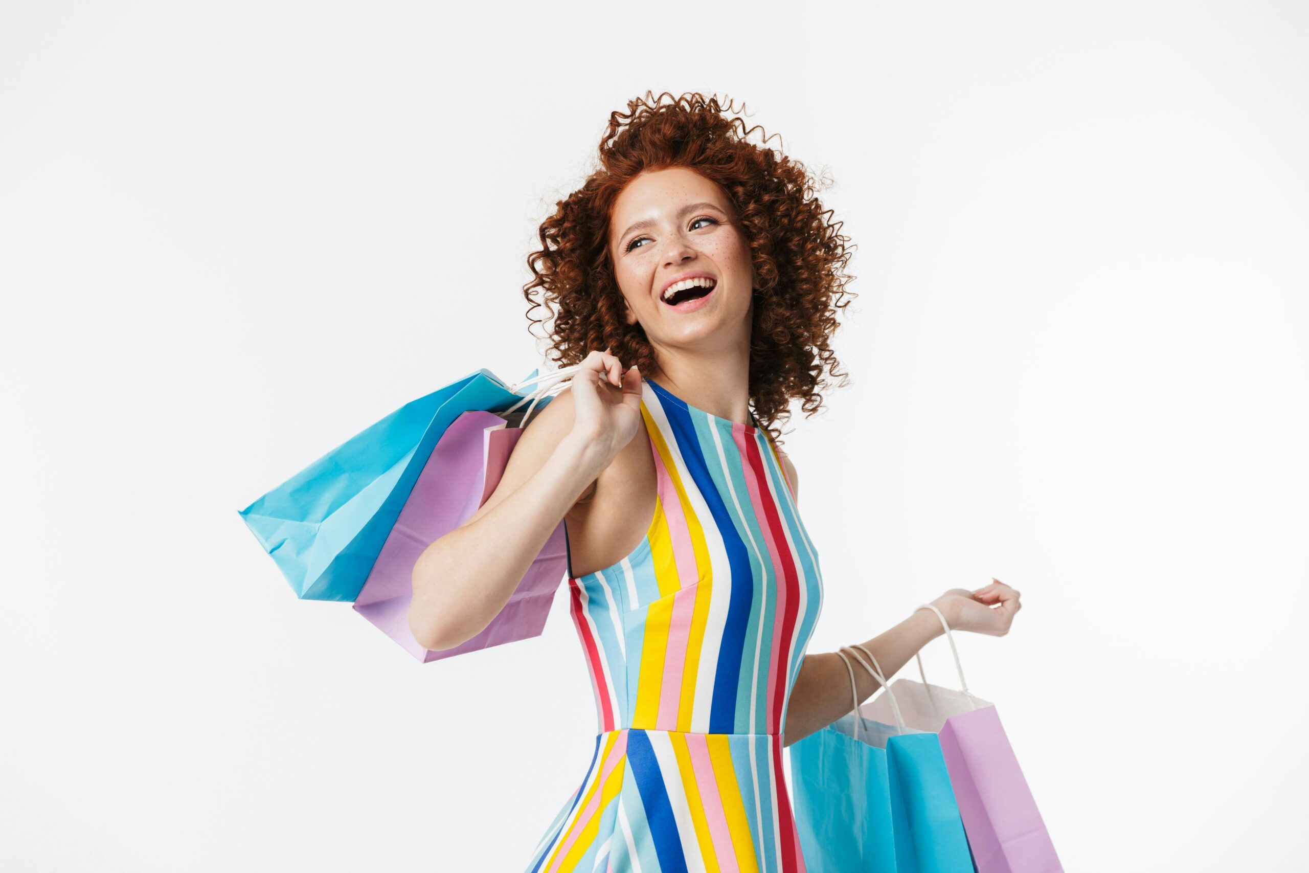 A cheerful young woman with curly red hair smiles while holding colorful shopping bags on a white background.