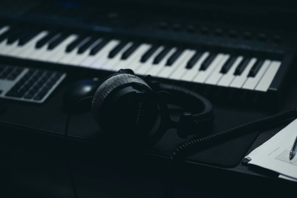 A pair of headphones sits on a dark desk in front of a musical keyboard in a dimly lit recording studio.