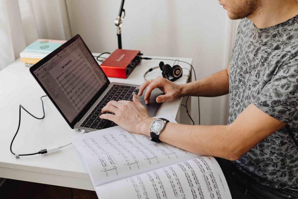 A side view of a musician working on a laptop with music software open, referencing printed sheet music on the desk.