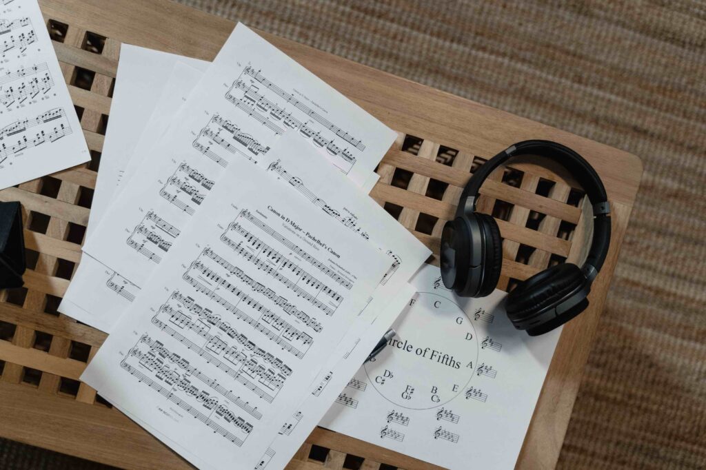 An overhead shot of printed sheets of music, including Pachelbel's Canon and a circle of fifths diagram, resting on a wooden lattice table next to a pair of black headphones.