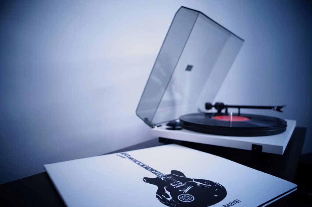 A vinyl record spins on a white turntable with an open dust cover, next to an album sleeve featuring a black electric guitar illustration.