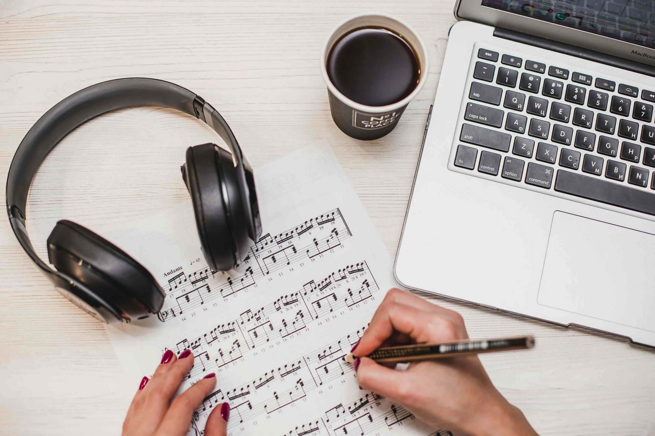 An overhead shot shows hands writing on sheet music next to black wireless headphones, a coffee cup, and an open laptop on a white wooden desk.