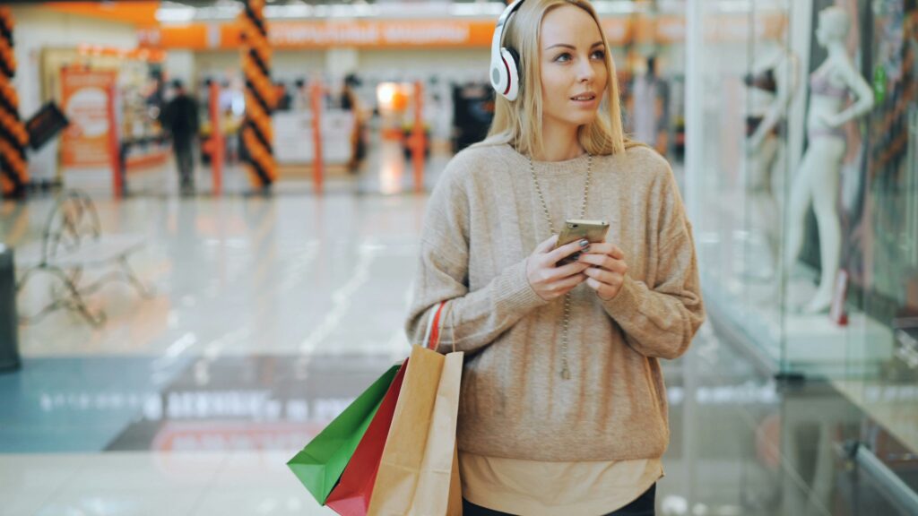 A young blonde woman wearing headphones and holding a smartphone walks through a shopping mall with paper shopping bags.
