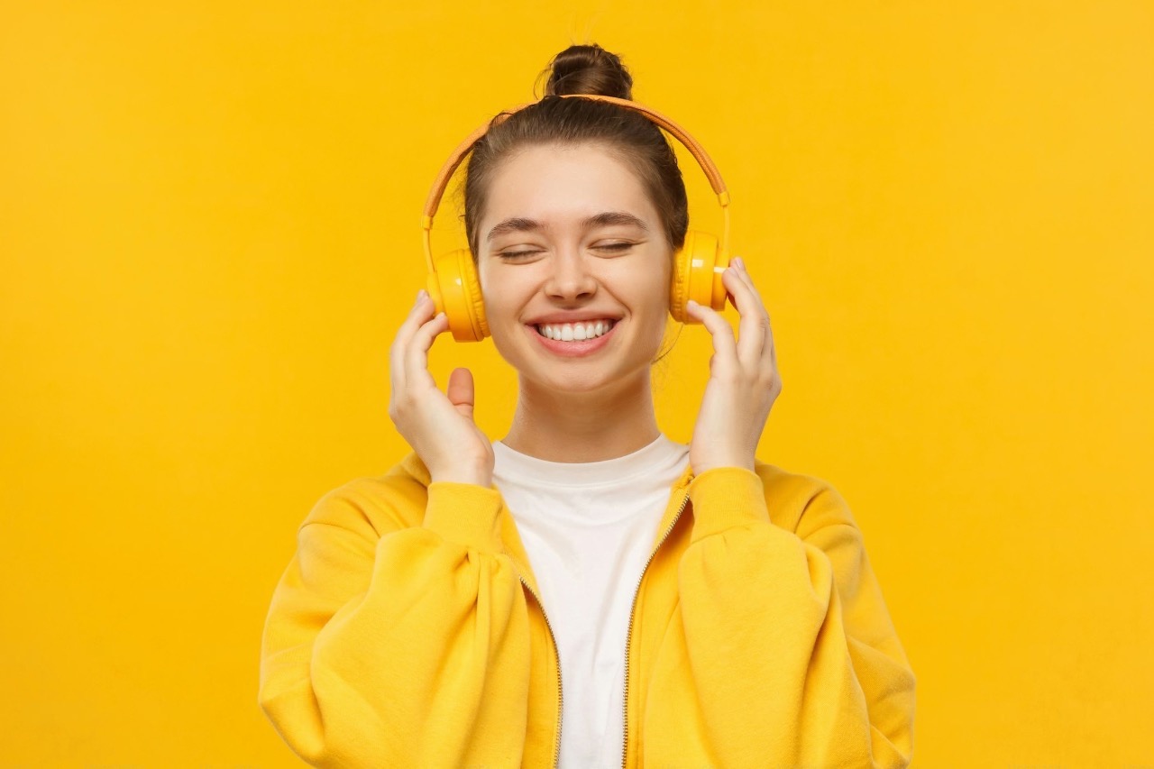 A smiling young woman wearing yellow headphones listening to music that improves mood against a bright yellow background.