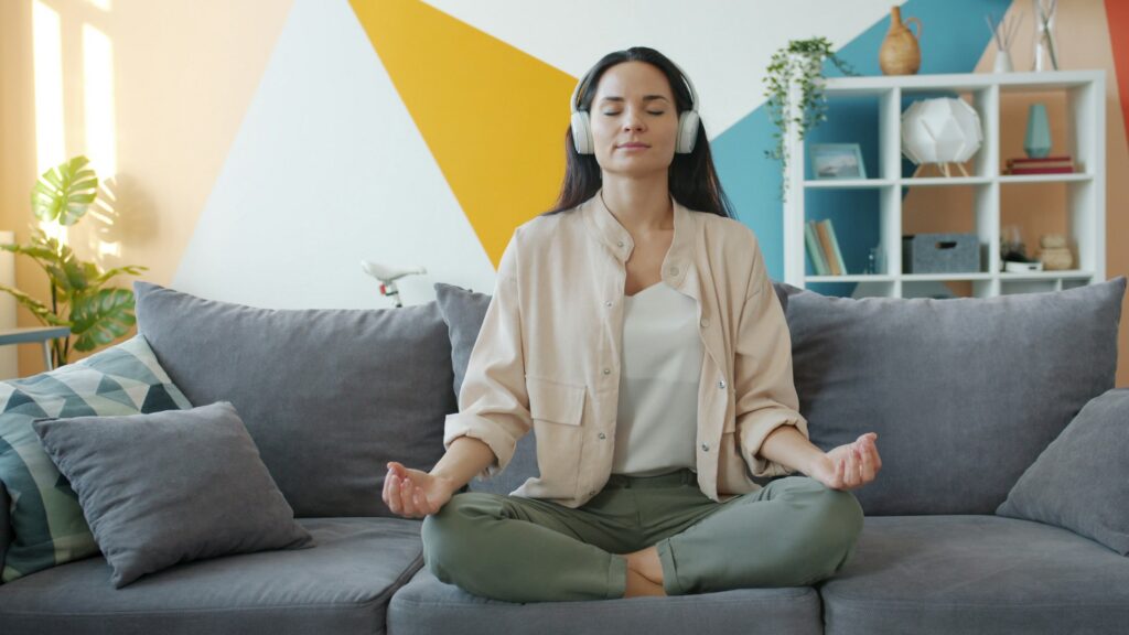 A young woman sitting in a lotus position on a sofa, practicing meditation while listening to relaxing music in wireless headphones to manage stress.