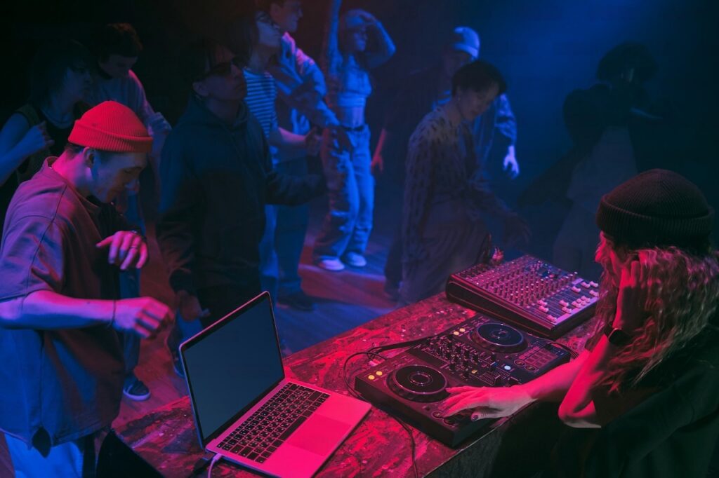 A DJ playing electronic music on a sound mixer at a club with young people dancing in the background under neon lights.