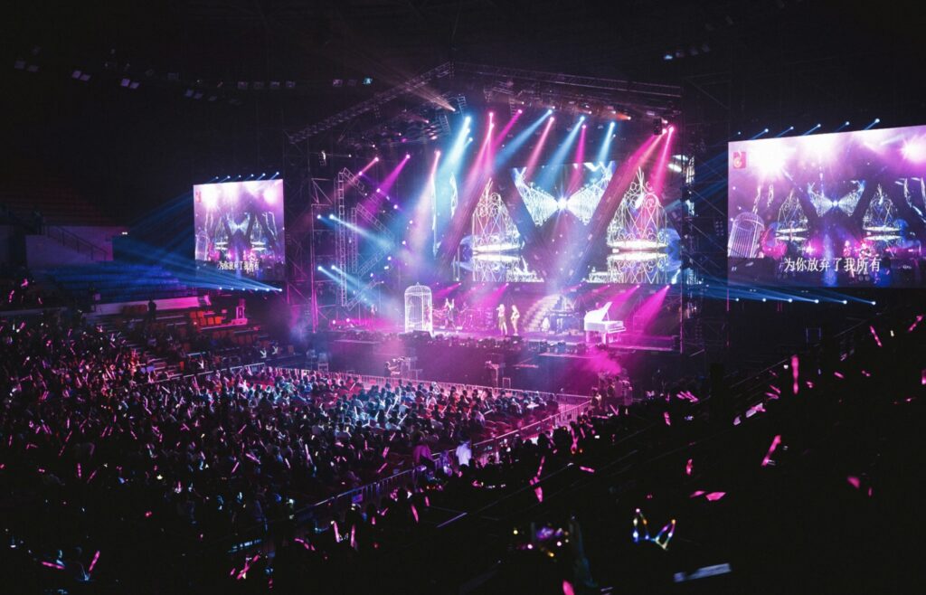 A massive crowd of fans waving pink lightsticks at a stadium K-Pop concert with a brightly lit stage and giant screens.