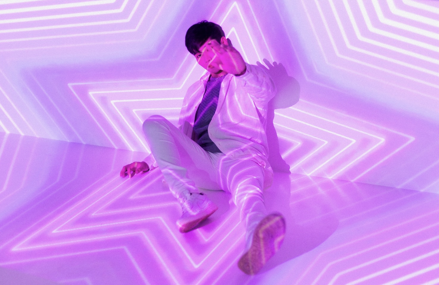 A young man in a white outfit sitting in front of neon purple star-shaped lights, representing the futuristic visual concepts of K-Pop music videos.