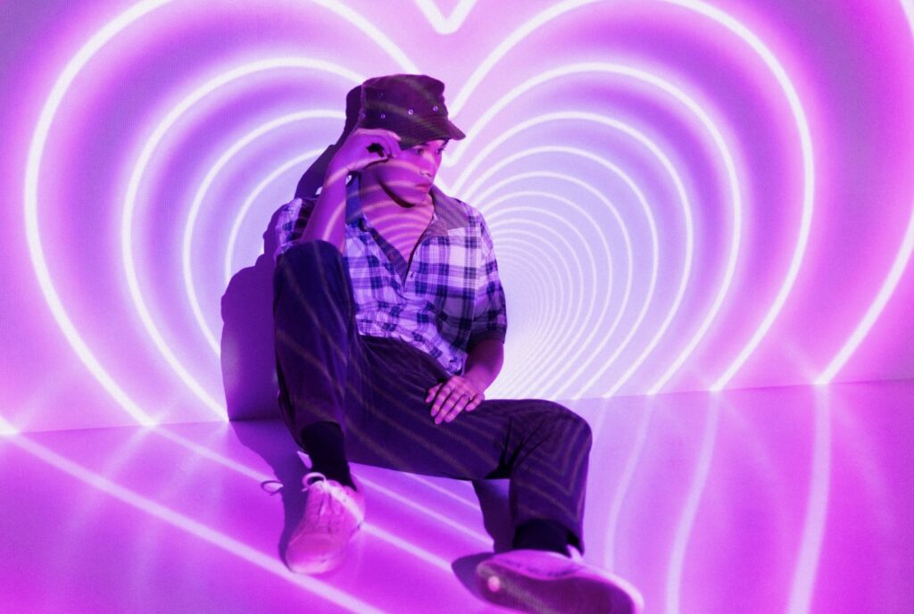 A stylish young man in a bucket hat sitting against neon pink heart-shaped lights, representing the visual pop concepts and idol aesthetics of K-Pop music.