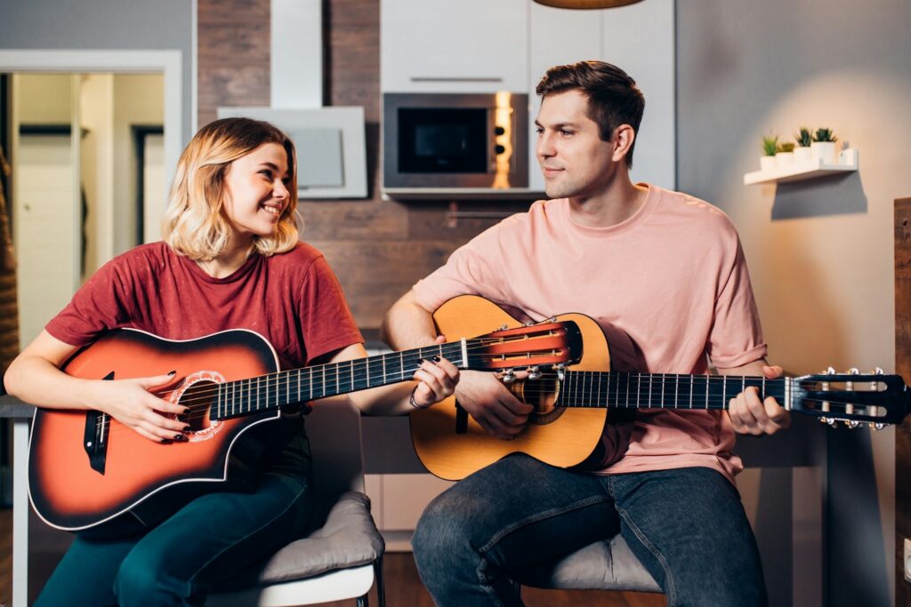 A young couple sitting in a kitchen and playing acoustic guitars together to experience social relatedness and positive emotional connection through music.