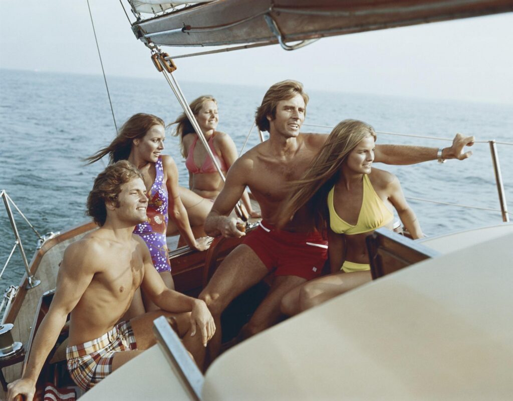 A group of friends in vintage swimsuits sailing on a luxury wooden boat, embodying the sun-drenched coastal escapism of yacht rock music.