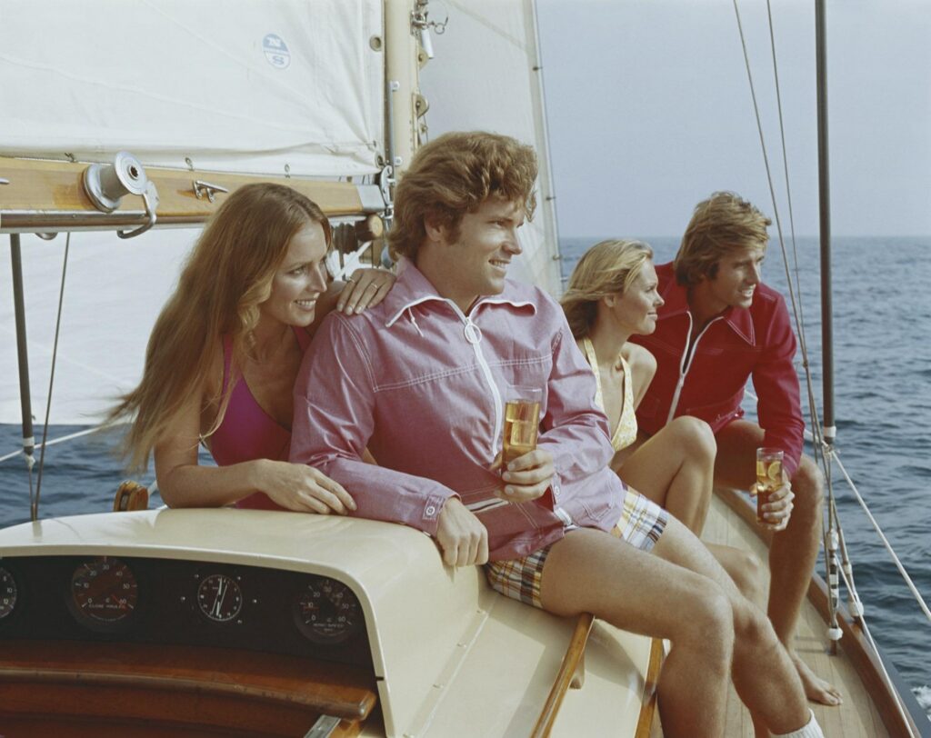Friends in retro 1970s coastal attire drinking cocktails on a luxury sailboat, representing the smooth and carefree yacht rock music vibe.